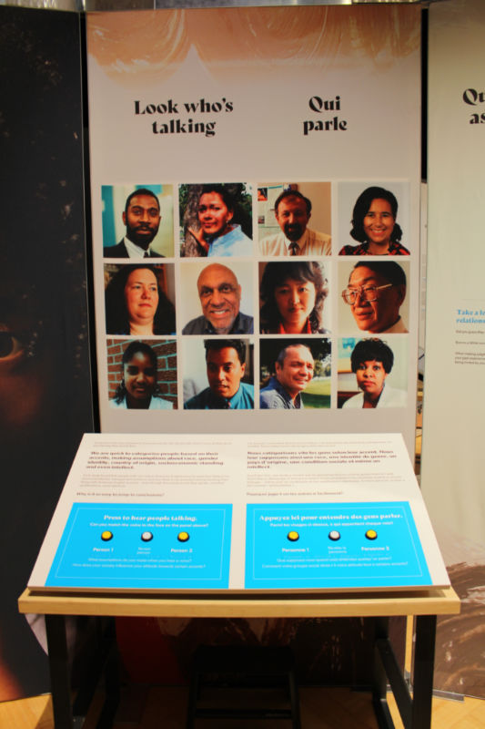 A museum display showing faces of 12 people with buttons on the table below to hear voices